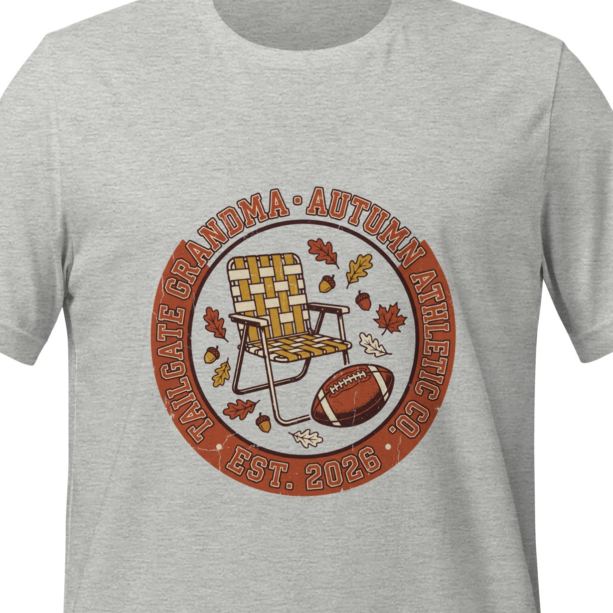 Athletic heather unisex t-shirt printed with a circular badge reading Tailgate Grandma and Autumn Athletic Co., arched around a folding lawn chair, a football and autumn leaves