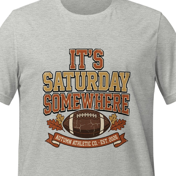 Athletic heather unisex t-shirt printed with bold stacked lettering reading It's Saturday Somewhere above a football and autumn leaves with a small banner beneath